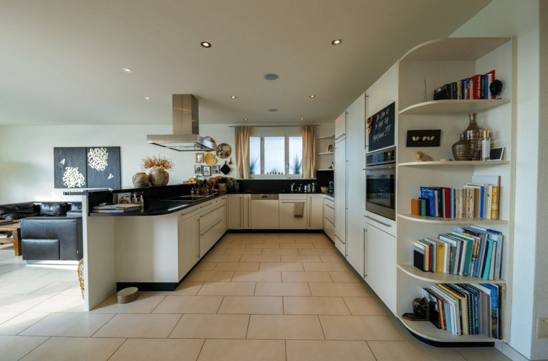 modern kitchen, with a center island, tiled floor, white cabinetry, open shelving with books, window with curtains, appliances including a stove and dishwasher, a fridge on the left, a sofa and a window in the distance