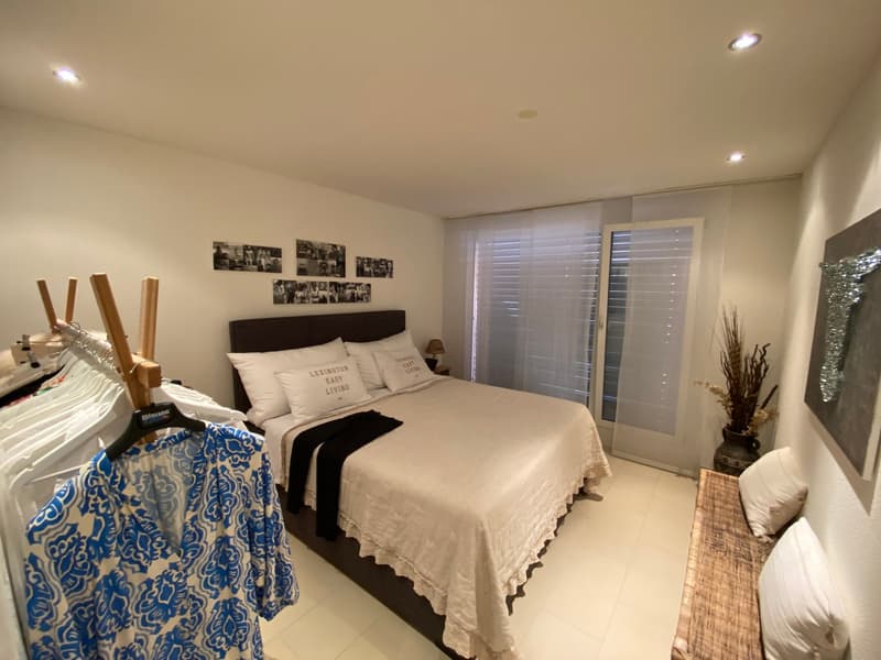 A well-lit bedroom with a white bed, bedheadboard with four white pillows, lamps on either side, a table with lamps, and picture frames on the wall. There is a cloth hanger, with some clothes hanging on it, on the left side. There is a bench against the w