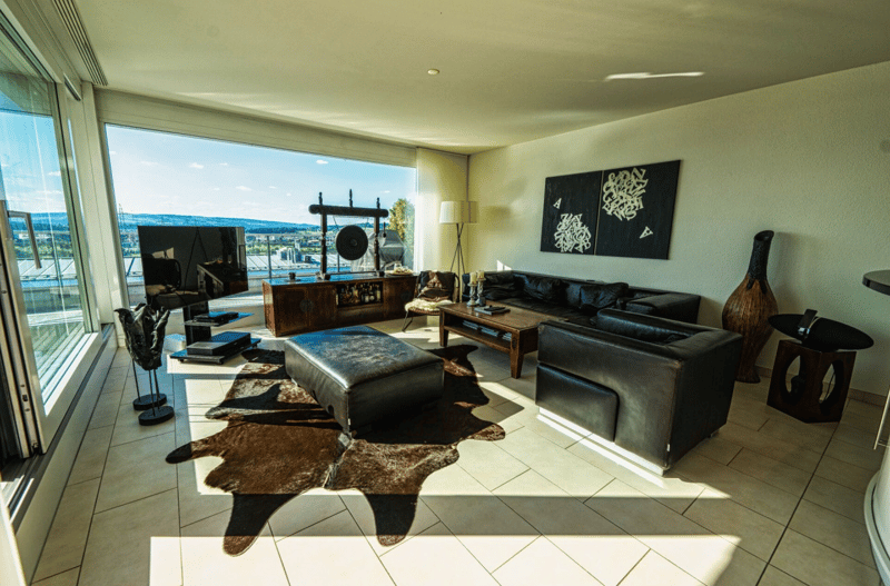 Open plan living room with large windows overlooking the city, large rug, black leather couch, coffee table, TV stand, decorative objects