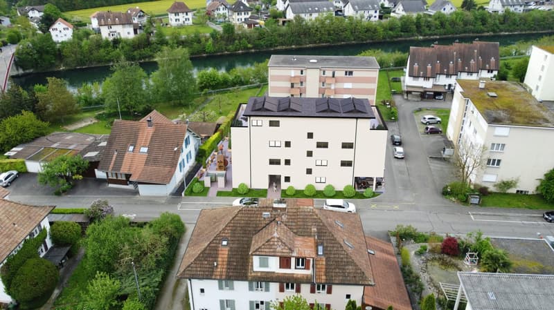 The image shows a residential area with a mix of multi-story apartment buildings and smaller houses. The buildings have a variety of architectural styles, with some featuring pitched roofs and others having flat roofs. The area is surrounded by lush green