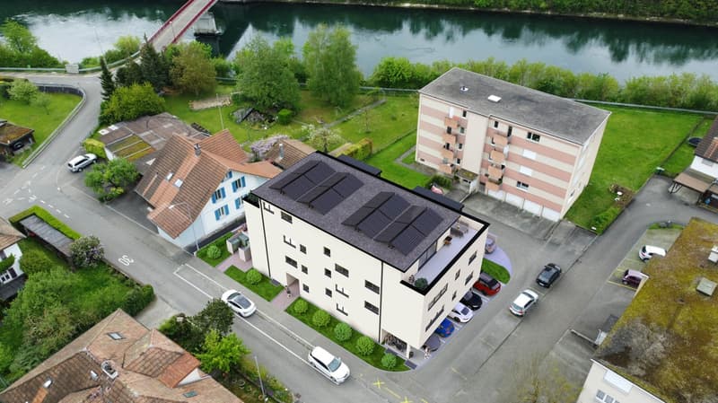 4-story apartment building with a flat roof, surrounded by trees and greenery, with a river or lake visible in the background. There is a parking lot in front of the building.