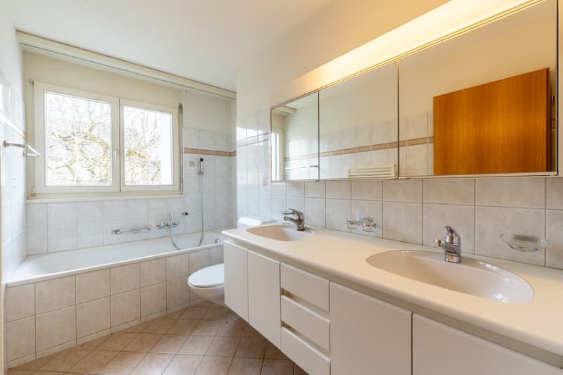 Bathroom with bathtub, two sinks, toilet, white tiles, wooden cabinet, window with view