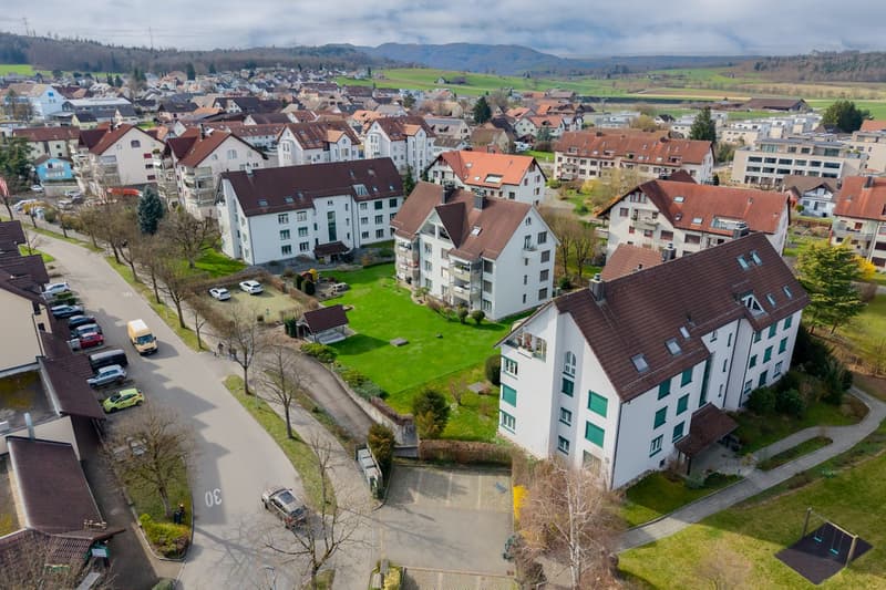 Aerial view of a residential area with multiple buildings, some with brown roofs, surrounded by green grass and trees, with a road and parking lot in the foreground.