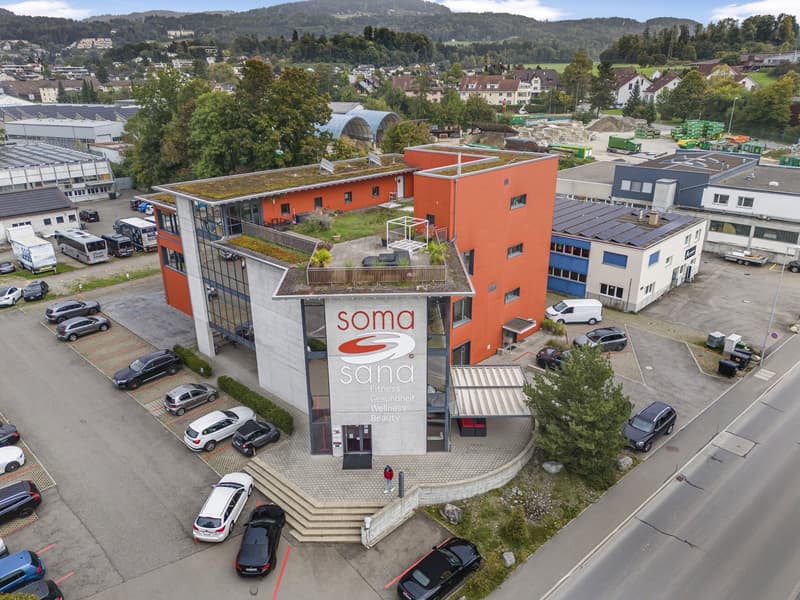 Red-orange commercial building with signage for 'SOMA SANA', multiple floors, parking lot with many cars, surrounding buildings