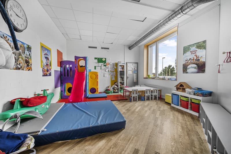 room with a clock, posters, colorful play toys, wooden flooring, storage shelves, windows, tables, chairs