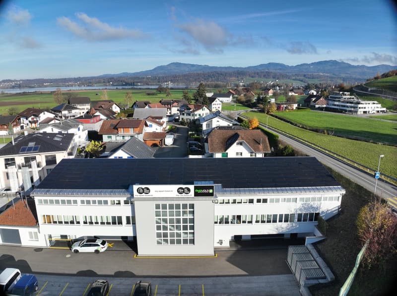 An aerial view of a modern white building with solar panels, a garage, a green field in the background, mountains and houses in the distance.