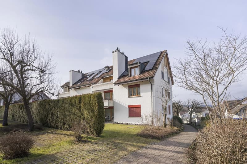 two-story white house with brown roof, solar panels on roof, small balcony on the first floor, well-kept garden, brick walkway