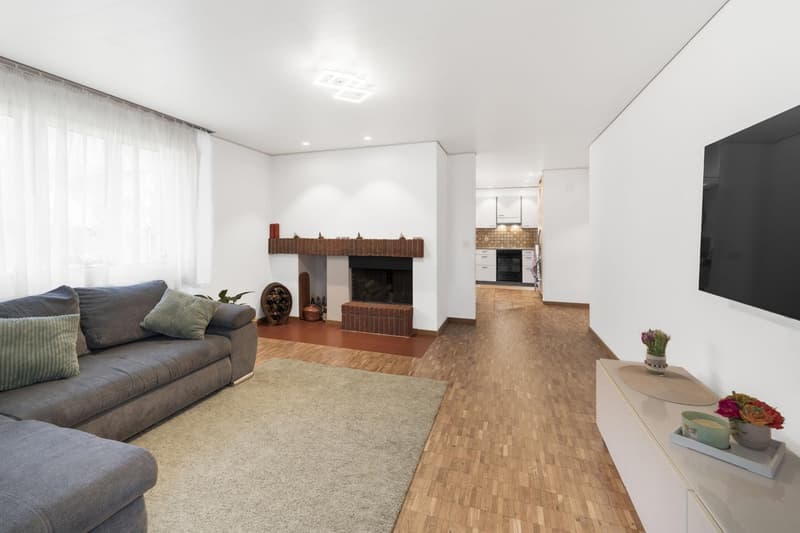 Living room with fireplace, gray couch, white walls, beige carpet, TV, decorative items, kitchen seen through archway