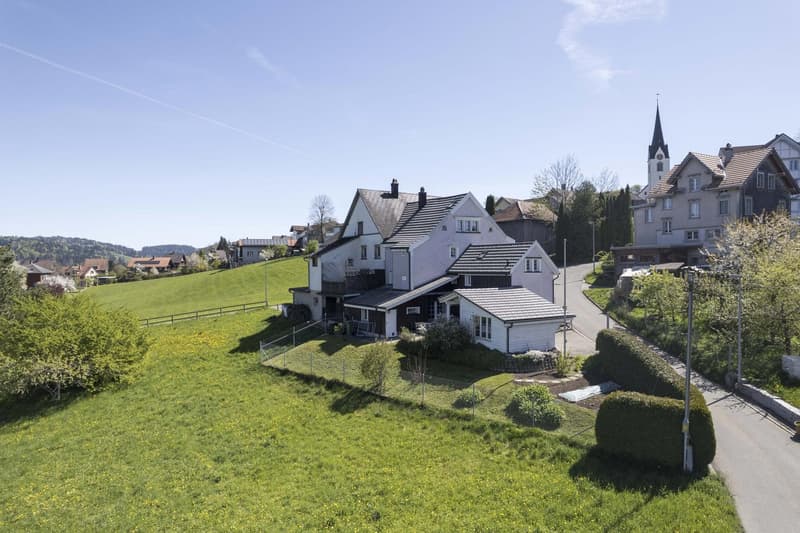 Aerial view of a large house with a gray roof, surrounded by a green field and a road. The house is located in a rural area with other houses and a church in the distance.