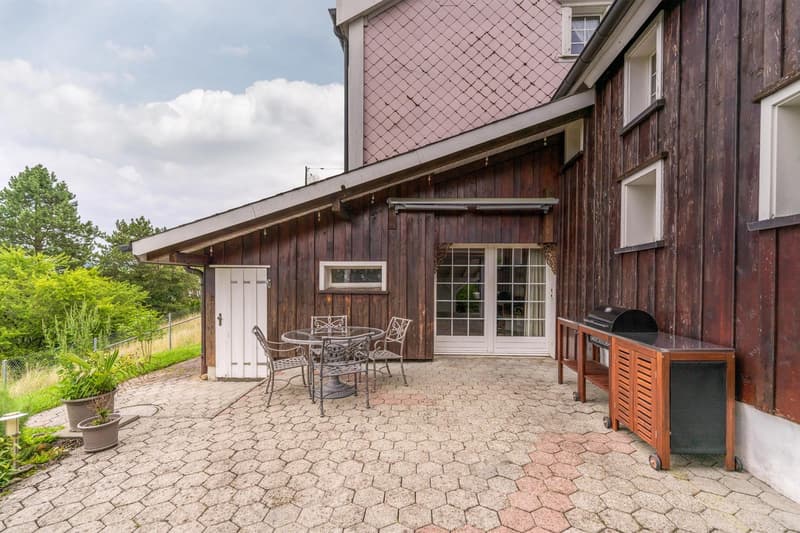 exterior with brown wooden siding, glass windows, white doors, paved patio, outdoor dining set, potted plants, grill station
