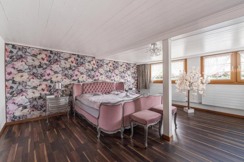 Pink velvet bed, wooden floor, floral wallpaper, white ceiling, nightstands, decorative pink pillows, and a pink stool