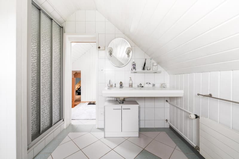 white tiled walls and floor, white vanity with double sinks, round mirror, shower door, shelf above sink, towel rack, radiator