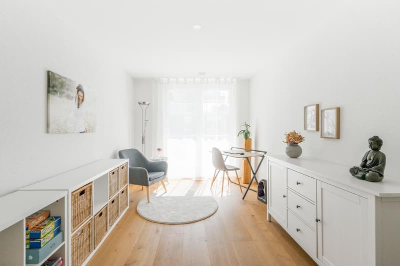 Minimalist room with white walls, wooden floor, white furniture, round rug, gray chair, desk, flower vase, framed pictures, Buddha statue, storage boxes, baskets, and a potted plant.