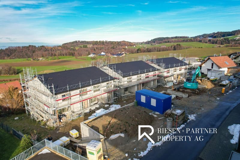 Aerial view of a construction site with several houses under construction. Some houses have metal scaffolding, others have roofing materials installed. In the background are a tractor and various construction vehicles. The site is surrounded by rural gree