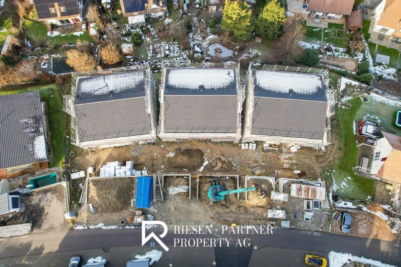 Row of houses under construction, with tiled roofs, some houses nearby in various stages of completion, snow on the ground, some plants, cars parked