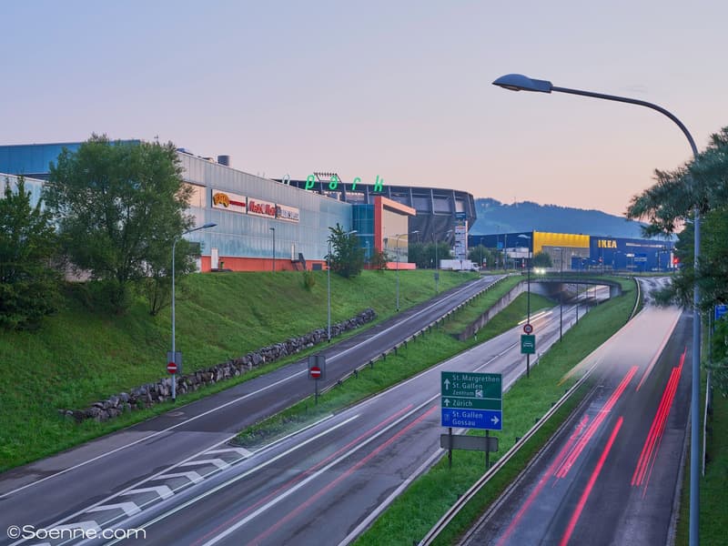 Highway with a busy traffic flow, streetlights on the side, road signs indicating directions and speed limits, lush green grass on the median and side slopes, buildings and commercial establishments in the background.