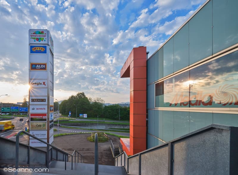 An outdoor view of a modern building complex with a large sign and outdoor signage, stairs, and a parking area.