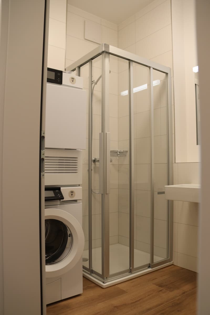 Modern bathroom with a shower stall, tiled walls, washing machine and dryer, and a white sink.