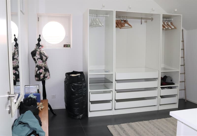 walk-in closet with white shelves, drawers, and hanging rods, a round window, a wooden table, a mirror, and a black trash bag