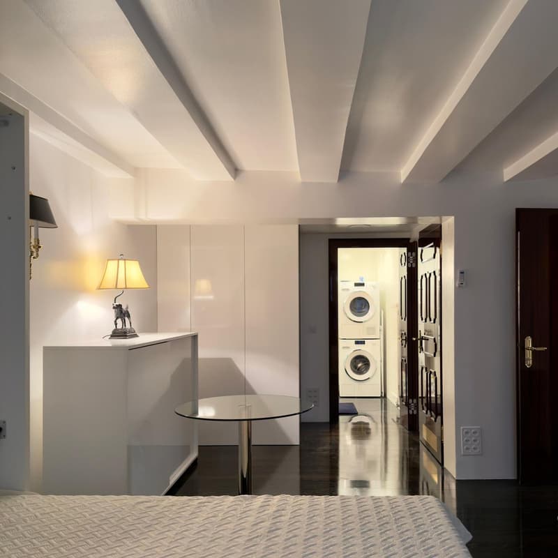 modern room with white walls, black floor, round table, lamp, white bed, white cabinets, washing machine