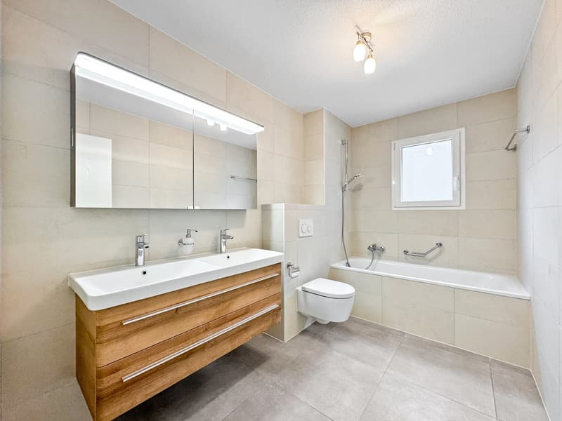 Modern bathroom with double sink, wooden cabinet, toilet, bathtub, and window.