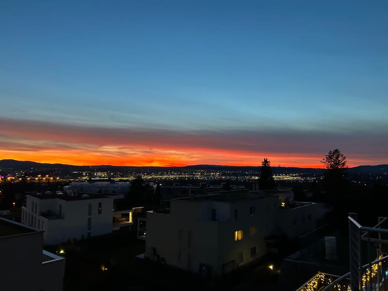 The image shows a scenic view of a city at sunset. The sky is filled with vibrant orange and red hues, creating a beautiful backdrop for the silhouetted buildings and trees in the foreground. The city lights are visible in the distance, adding to the over