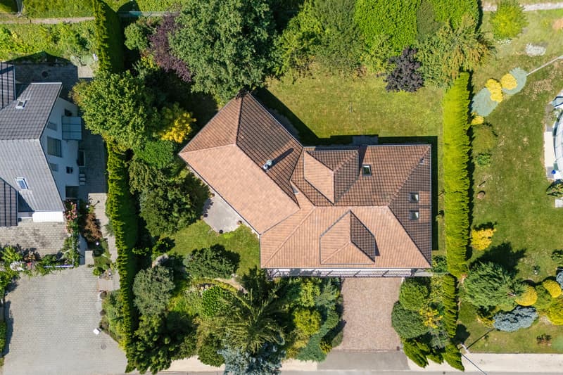 Aerial view of a house with a brown roof, surrounded by trees and grass, driveway, and a neighboring house