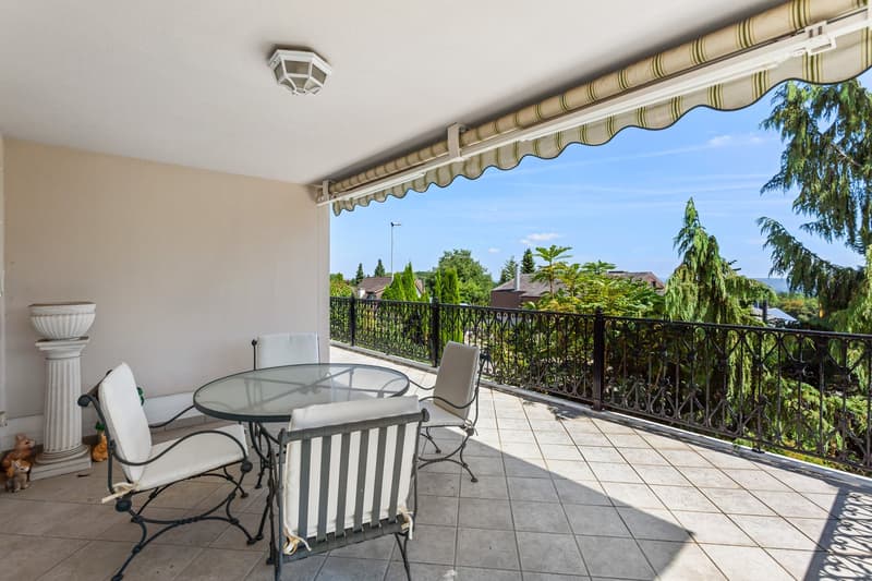 Balcony with table and chairs, black railing, tiled floor, outdoor lighting, view of trees and houses