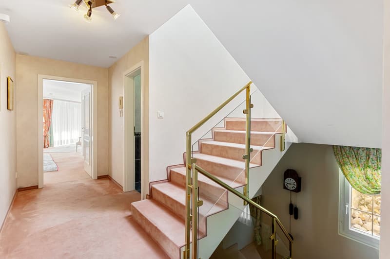 Staircase with pink carpeted steps, gold railings, white walls, hallway with wooden floor, picture frames, and a clock on the wall.
