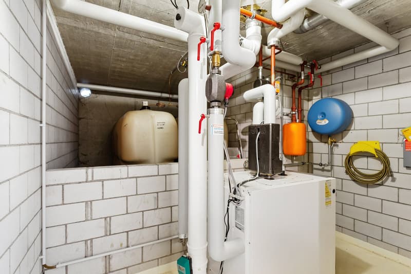 Utility room with white brick walls, white pipes, white and orange tanks, white washing machine, yellow hose holder, and a light on the ceiling.