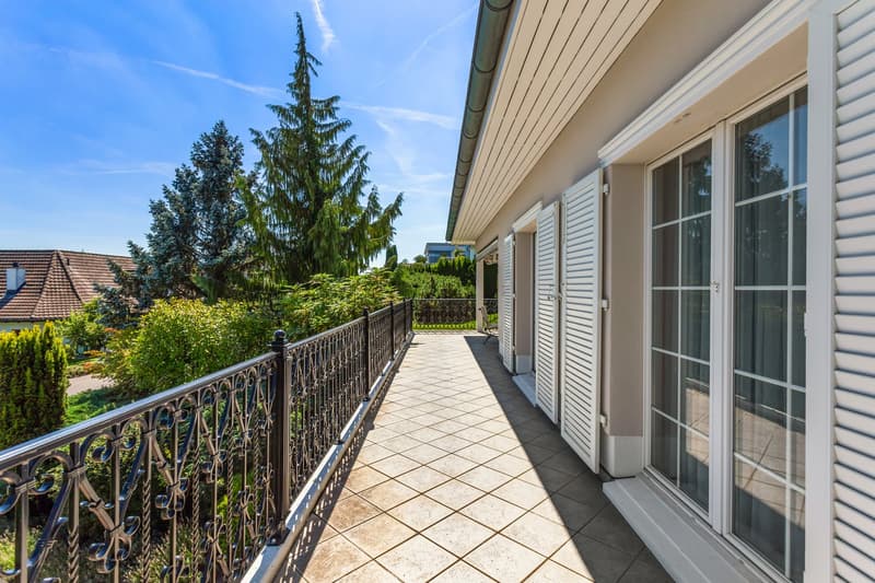 Balcony with tiled floor, black railing, white shutters, glass doors, view of trees and houses
