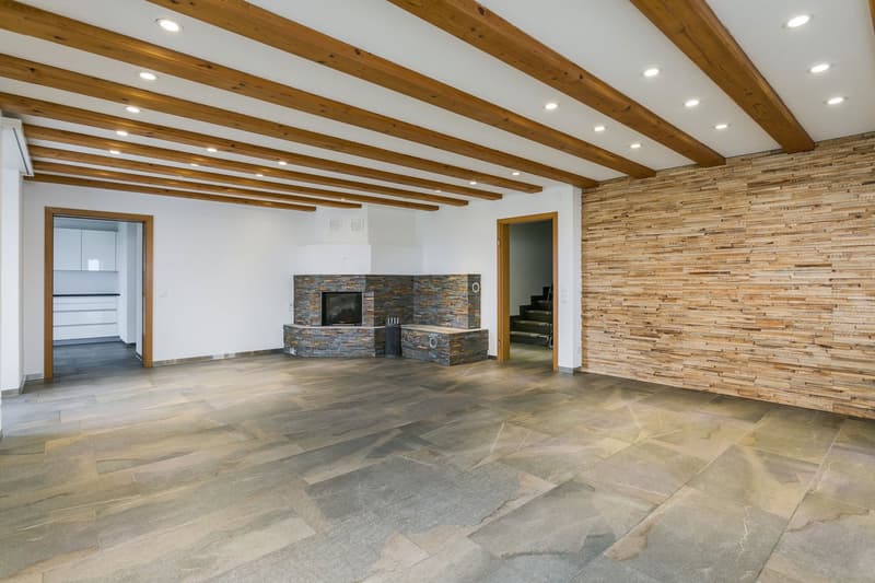 Large open-concept living space with exposed wooden beams on the ceiling, a stone fireplace, and a tiled floor with a natural stone pattern. The room has a modern and rustic design aesthetic.