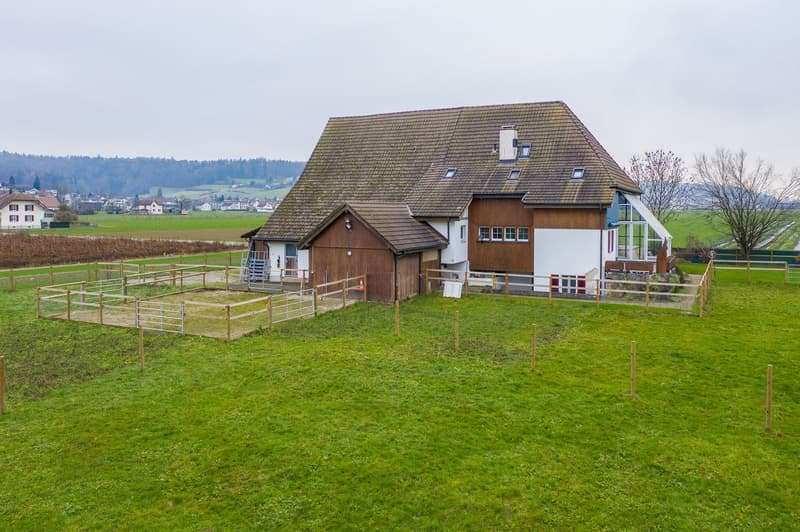 2-story house with a wooden exterior, a tiled roof, and dormer windows. The house has a garage and a balcony or terrace. It is surrounded by a grassy field and fenced enclosure, with a rural landscape in the background.