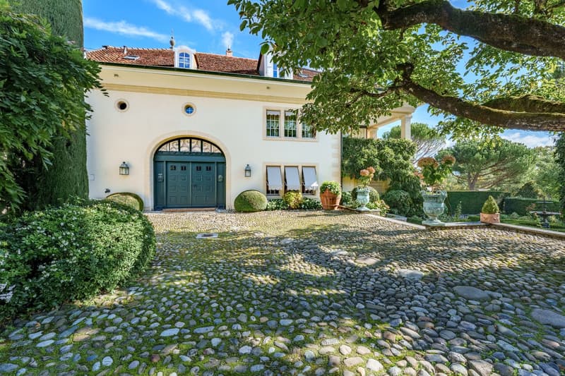 White building, red roof, double front door, green door, gravel driveway, large tree, plants and shrubs, lamp on both sides of the door
