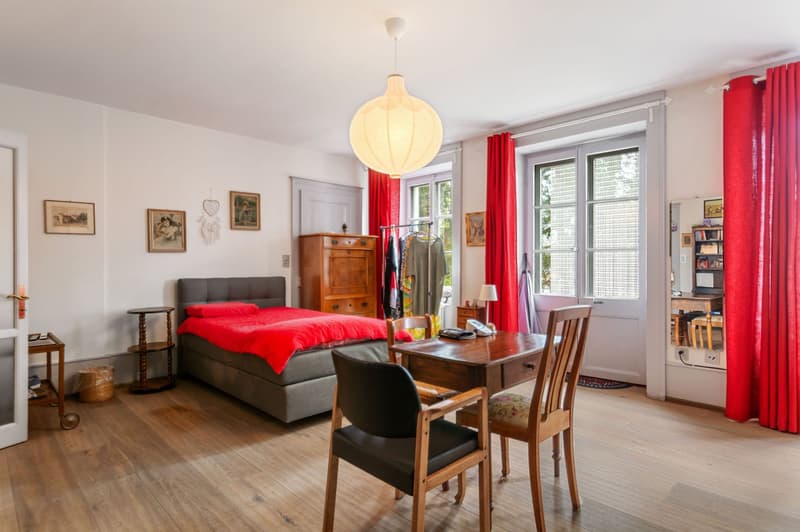 bedroom with bed, two chairs, desk, red curtains, wooden floors, pendant lamp, wall decor, telephone