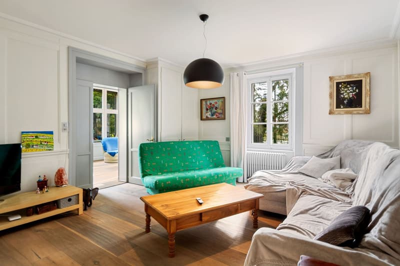 Living room with wooden floor, white walls, large windows, couch, sofa, wooden coffee table, remote control, television, artwork, wooden television stand, and an elephant sculpture