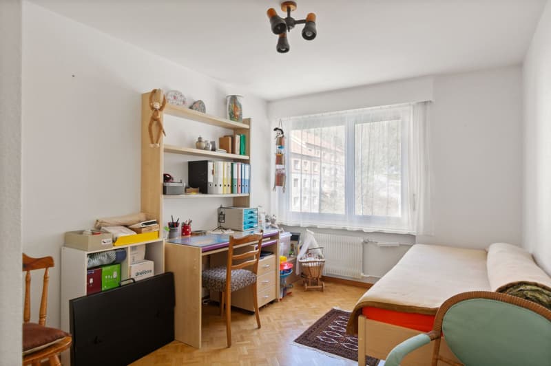 desk with chair, bed, bookshelf with items, floor radiator, window with curtains, rug, wicker basket, children's bed