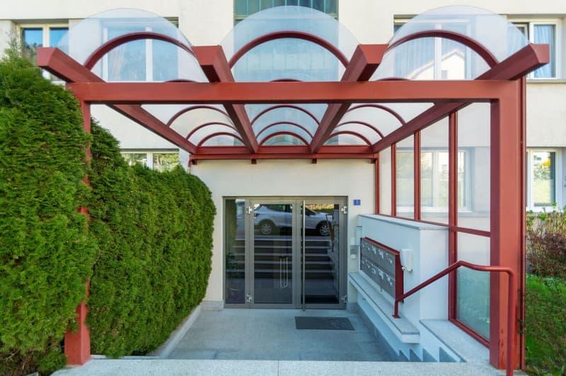 Modern apartment building entrance, glass doors, red metal structure, steps, mailbox, parking