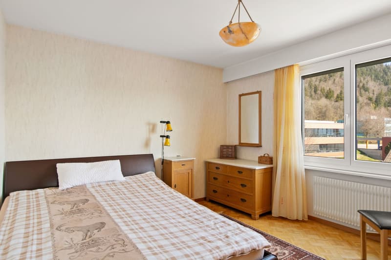 bedroom with a double bed, a wooden dresser, a nightstand, a lamp, a radiator, a mirror on the wall, and a window with curtains