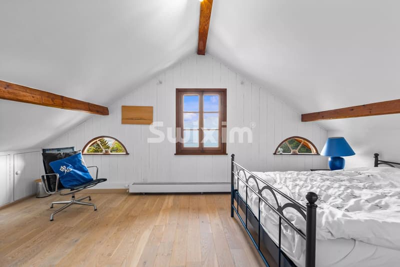 Une chambre lumineuse avec poutres apparentes, un lit en métal, une lampe bleue et une chaise moderne, offrant une vue par la fenêtre centrale.