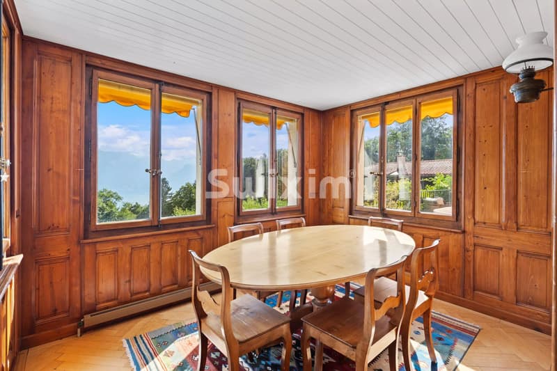 Une salle à manger avec un mobilier en bois, des fenêtres donnant sur l'extérieur et un tapis coloré sous la table.