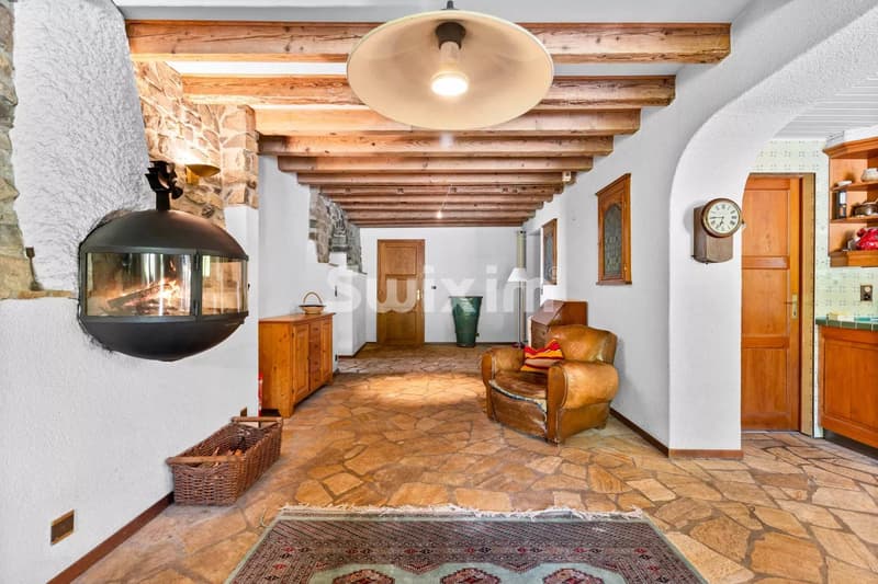 Une pièce chaleureuse avec un plafond en poutres apparentes, un sol en pierres, et un fauteuil en cuir près d'une cheminée moderne sur le mur.