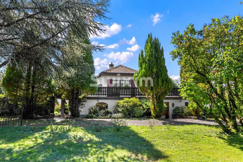 Grande maison entourée d'arbres avec un jardin verdoyant sous un ciel bleu.