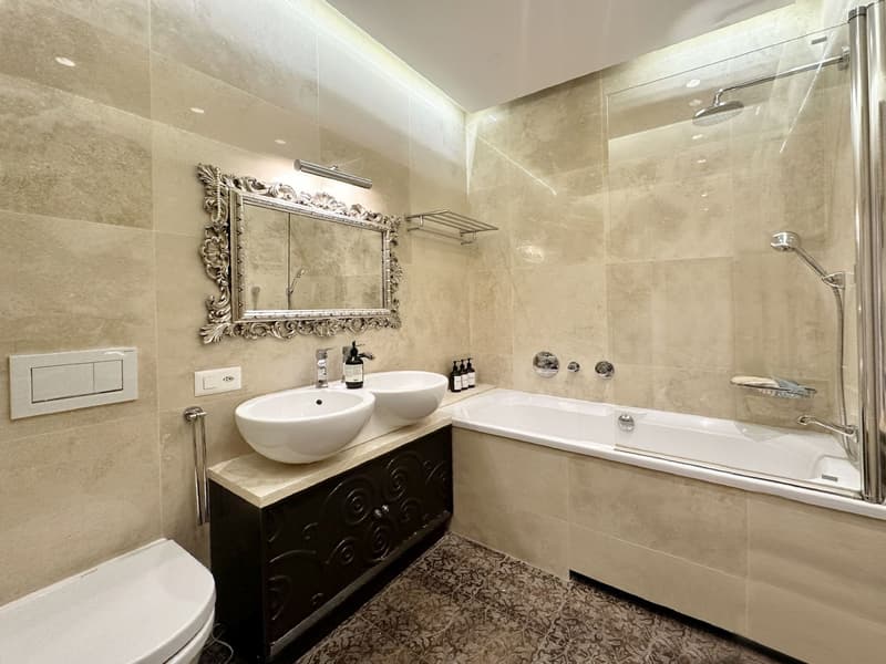 Bathroom with white marble tiles, white sink, bathtub, shower enclosure, decorative mirror, and modern fixtures.