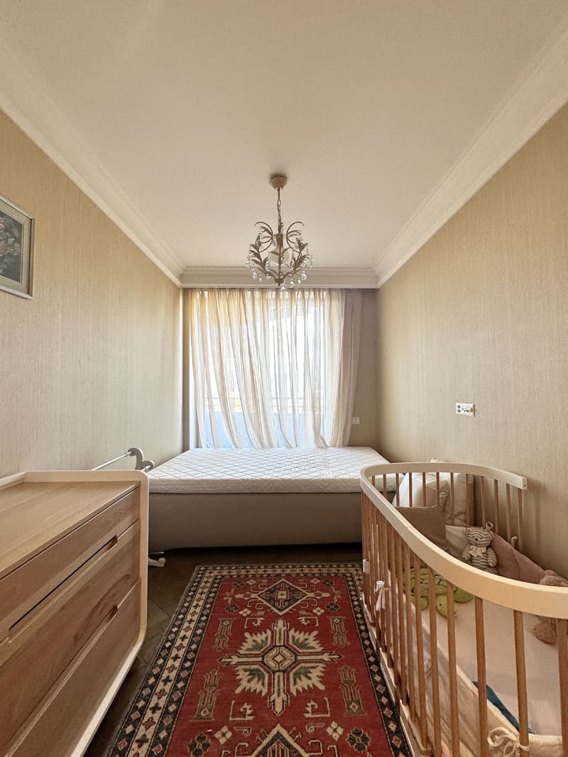 Bedroom with bed, chandelier, crib, window with curtains, wooden dresser, oriental rug