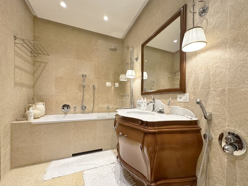 Spacious bathroom with bathtub, vanity, and separate shower area. Marble flooring and tiled walls. Wall-mounted towel rack and built-in mirror.