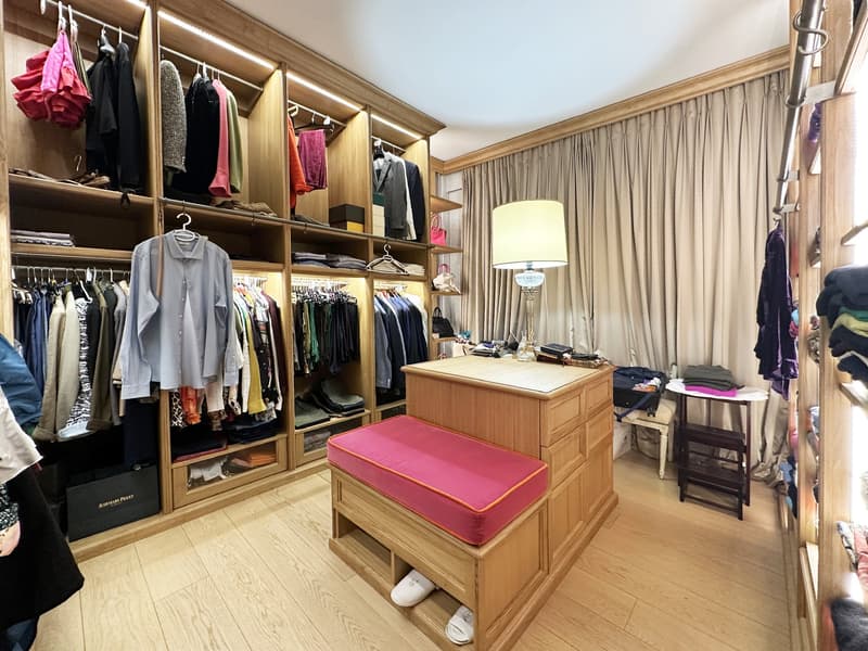 walk-in closet with wooden shelves, clothing items, pink cushion bench, wooden floor, white shoes, table, lamp, curtains
