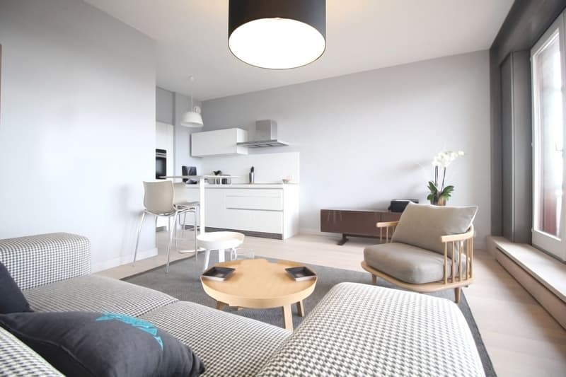 modern living room with white walls, wooden floor, gray couch, wooden coffee table, white kitchen, dining table, chairs, white ceiling lamp, white flower vase