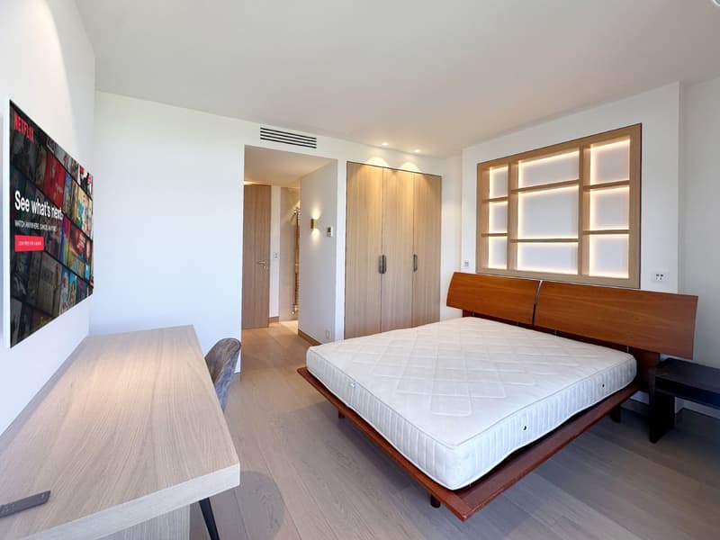 Bedroom with wooden bed, wooden headboard, wooden desk, wooden chair, wooden closet, white walls, wooden floor, wall-mounted TV, and a poster on the wall.