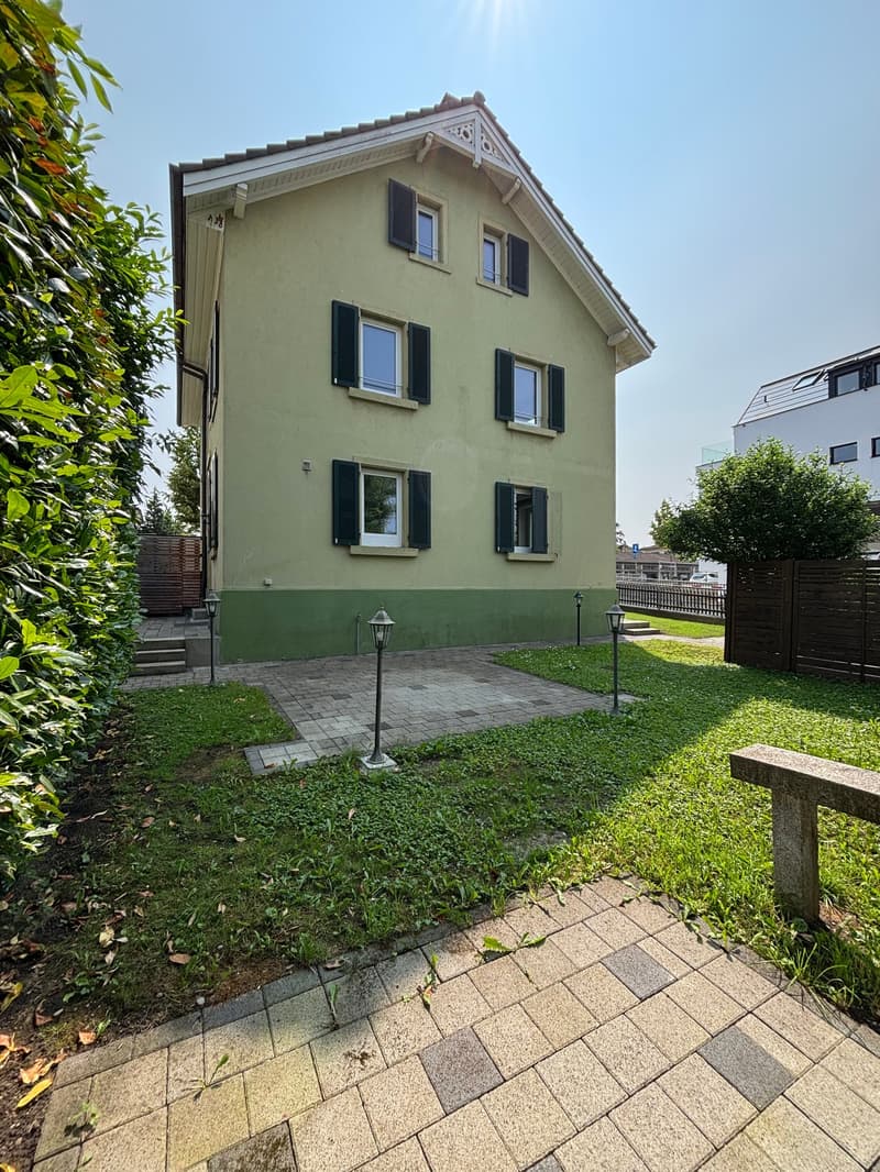 2-story house, light green facade, black shutters, brick walkway, front yard with grass and plants, 3 lamp posts, small wooden fence